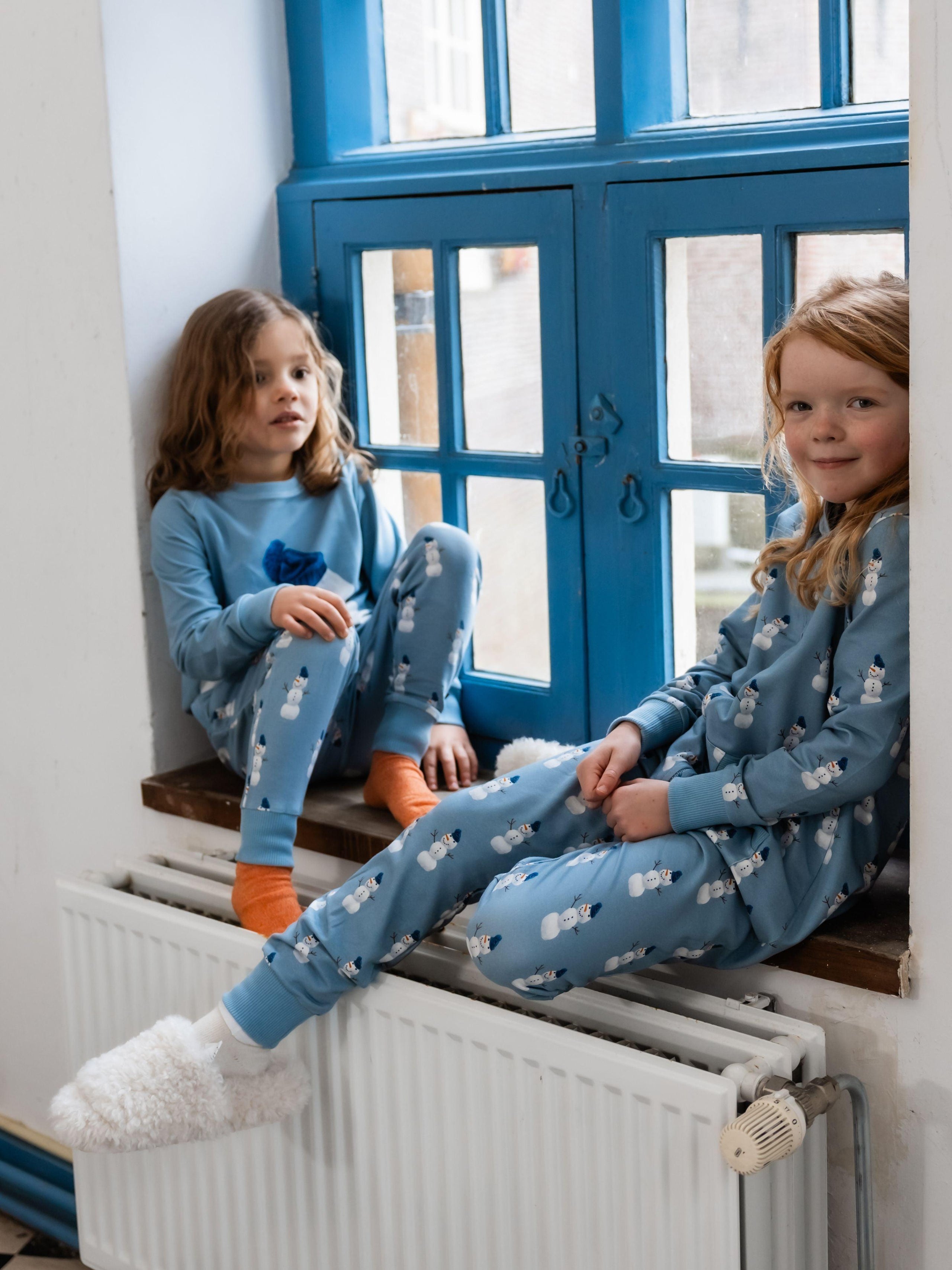 Kids wearing blue pajamas with a snowman print in a blue windowsill